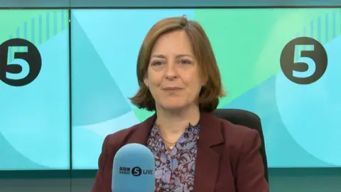 Dame Melanie Dawes is looking at the camera in a 5 Live studio, there is a microphone in front of her. She has chin length hair and is wearing a maroon suit jacket and a blue and maroon shirt.