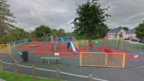 Google Maps street view of a playground in Odd Down, Bath. There is a swing set, a slide and roundabout as well as two green benches and a bin. A housing estate can be seen behind it. 