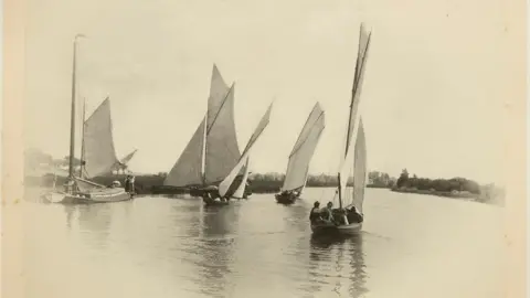 Sepia photo of boats being sailed on the Norfolk broads. There are four vessels each with a sail. You can see there are three people in the boat closest to the camera. 