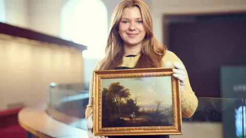Cimbalo family/Lyon & Turnbull Amelia Cimbalo has long blonde hair. She is wearing a yellow jumper and protective gloves as she holds the painting. Amelia is standing in a large, well-lit space.