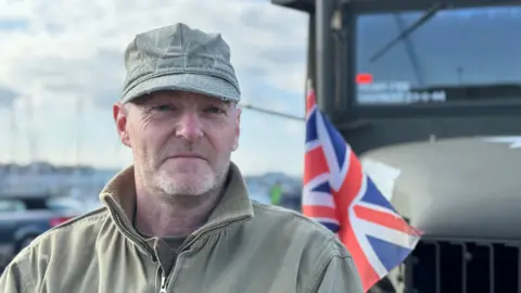 Mr Bagely, who is wearing military-style khaki clothes, is stood in front of a large green truck with a Union Jack flag.