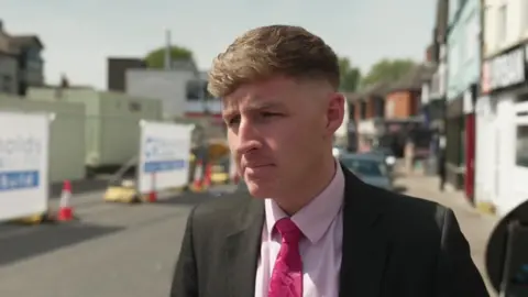 A man wearing a black suit, with a pink shirt and bright pink tie, stands in front of a building project on a town centre street on a sunny day.