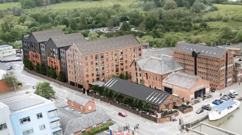 A computer-generated image of a six storey, warehouse-style building behind Alexandra Warehouse, near the Gloucester and Sharpness Canal. There is green space with trees behind the proposed development.