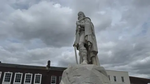 The statue of King Alfred is pictured on a plinth and taken from the ground nearby. A roof is visible in the background the skies are grey and overcast. 