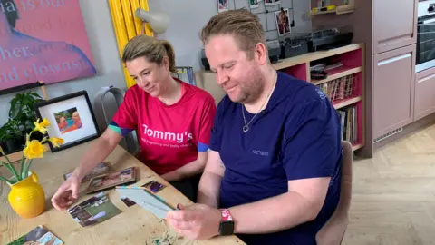 Ben Jelley sits at the kitchen table looking at photos of his wife with family friend Emily Mayhew. She is running the London Marathon with him and is wearing a pink Tommy's t-shirt.
