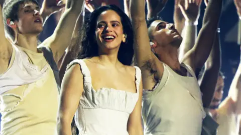 Spanish singer Rosalia on stage in a white dress laughing and looking to the left - some backing dancers can be seen behind 