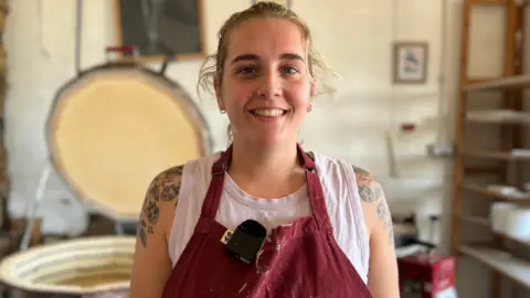 A woman wearing burgundy dungarees in a pottery studio.