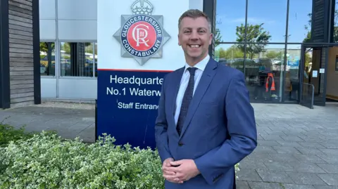 Nick Evans, a man perhaps in his 50s with a navy suit, pale shirt, and tie, smiles with his hands held together as he stands in front of the Gloucestershire Police headquarters sign at the front of the building.