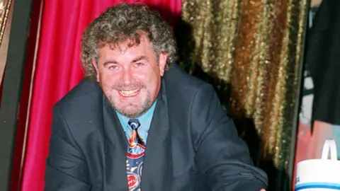 Gerry Kelly is smiling broadly at the camera. He has curly brown hair which is greying slightly. He's wearing a dark suit, with a blue shirt and a brightly coloured tie