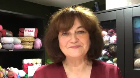 Darren Rozier/BBC A general view of Sharon Cane, owner of the Knitters Attic. She is standing in the shop and smiling to the camera. She has shoulder length brown hair and is wearing a dark red dress. Behind her are black shelves which have tens of yarn balls placed upon then.