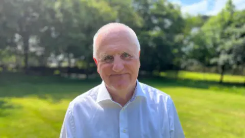 BBC Deputy Peter Ferbrache stood in his garden wearing a white shirt. He has very short white hair and is clean shaven. 