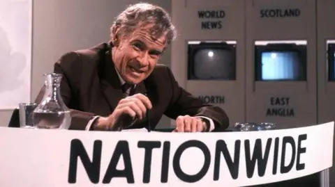 Baxter pretending to be a Nationwide presenter for a sketch. He is sitting at a desk with a huge sign that says Nationwide in front of him.