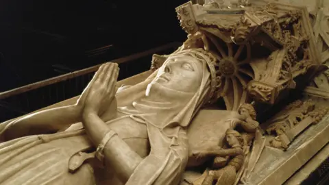 Hulton Archive via Getty Images A tomb made of alabaster is carved to show a woman lying with her hands in prayer. She has a pillow under her head with miniature men plumping it for her comfort.