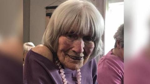 Hilda Bond smiling at the camera, she appears to be in the care home as there are other elderly residents around her. She is wearing a purple jumper with a pink beaded necklace around her neck. Her hair has been styled and lays flat and she has a fringe
