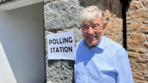 An elderly white man with grey hair in smart blue shirt smiling at camera in front of a polling station sign.