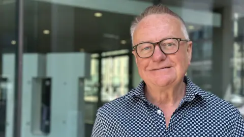 Professor Paddy Gray is wearing glasses and a blue checked shirt. He has short grey hair and he is smiling at the camera. Professor Gray is standing in front of a window at Ulster University's campus in Belfast.
