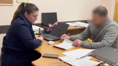 A police officer in blue uniform sits opposite a man in a grey sweater and blurred face at a table. The man is reading a sheet of paper, with a pen held in his right hand. Other pieces of paper are scattered on the table, where a mobile phone and charging lead, two laptops and thick lever-arch file can be seen. 