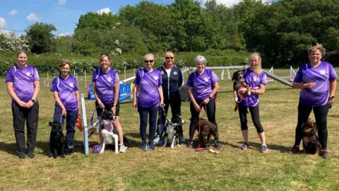 The seven competitors are pictured wearing their purple club tops. Six of the women have their small and medium sized dogs with them. They are stood in an agility field with the apparatus visible behind them.