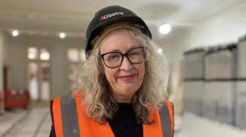 Shaun Whitmore/BBC Woman with long blonde/grey hair wearing spectacles, an orange hi-vis vest and a black hard hat with Gipping written across the front