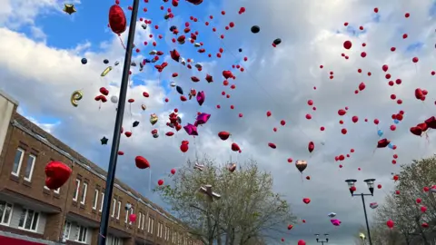 Lots of balloons, which are mostly red or pink, rise up to a cloudy sky having been let off by people below.