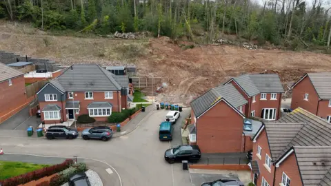 Elevated view captured by a drone of the bank at Haden Cross Drive, with soil blocking the road below and about six houses in the foreground, with cars parked on the road