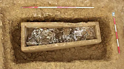 A dug-up grave with a stone coffin inside it and red and white markers placed around the grave.