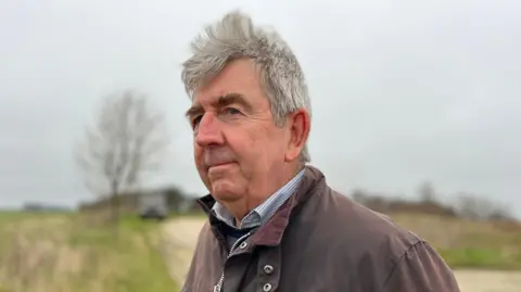 Norman Beale standing outdoors. He is wearing a brown waxed coat with a blue shirt and jumper beneath the coat and he is looking into the distance. He has grey hair which is blowing in the wind. There is a path behind him with grass on either side.