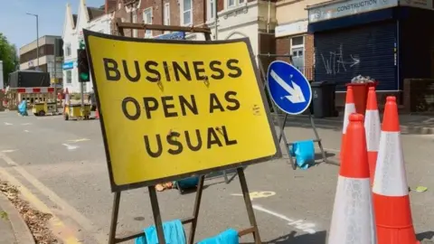 Roadworks on a road with a bright yellow sign saying Business Open as Usual.