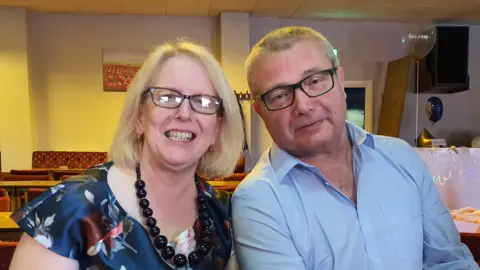 Handout A family photograph showing Audrey Buckham, left, wearing a black necklace along with a floral dress, smiling while next to her late husband, Eddie, who is wearing black rimmed glasses and a blue buttoned shirt. They are both inside a function room.