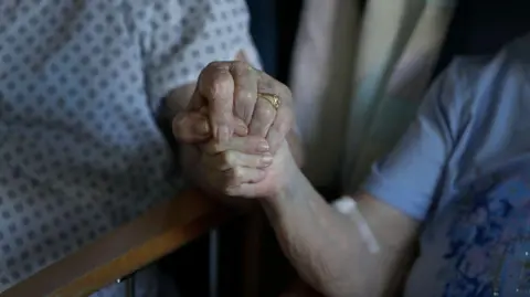 Close up of an elderly couple holding hands. One person wears a hospital gowns and a gold ring. The other is wearing a purple t-shirt. Their faces are not in view. 