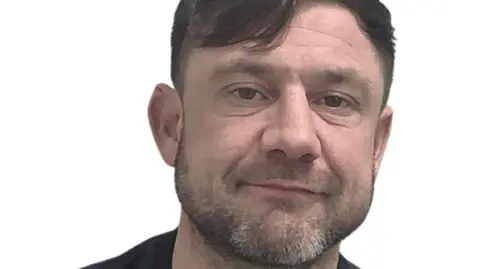 Custody image of Scott Warner, a man with a dark brown floppy fringe in a black t-shirt has a slight smile as he looks directly at the camera