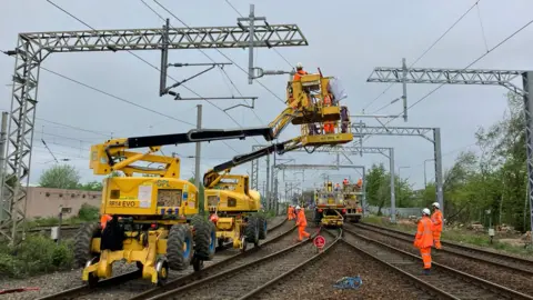 Network Rail People in orange high vis jackets and trousers stand along a railways line. Four yellow cherry pickers are raised up to the railways power lines, people in the baskets are working on the lines.