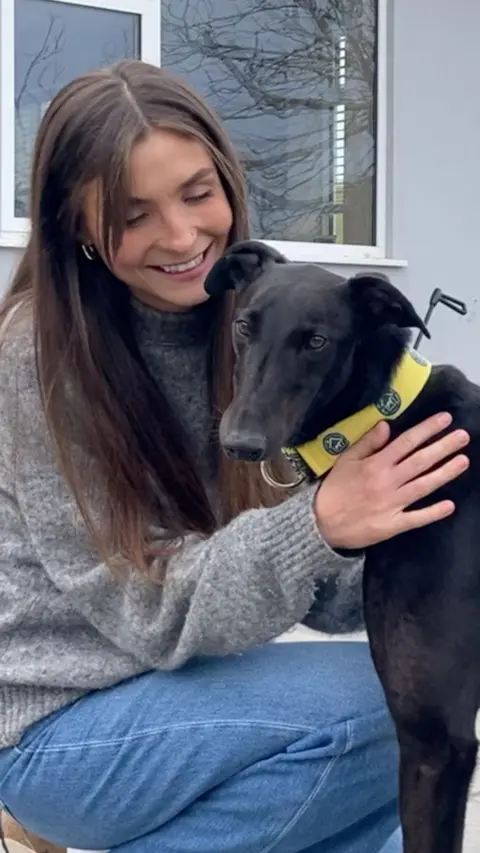 Woman with long brown hair wearing a grey jumper and blue denim jeans with a black greyhound wearing a yellow collar