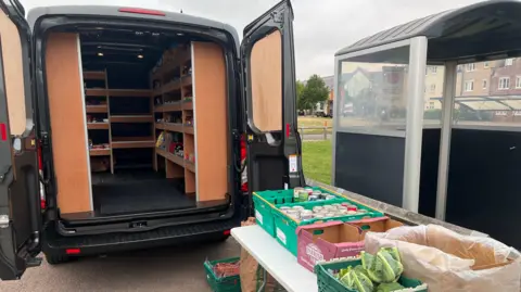 DJ McLaren/BBC The back of a van with doors open showing shelves full of goods and behind it a table with crates of tinned and fresh vegetables