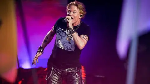 A rock singer on the Pyramid Stage at Glastonbury. He has reddish brown hair and is wearing black leather trousers and bright purple lights illuminate the stage. He has tattoos on both of his arms and is singing into a microphone.