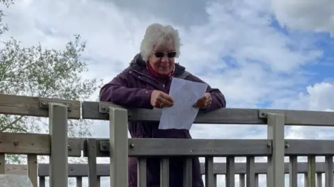 Doreen Savage Mrs Savage rests over a bridge looking at a piece of paper. She has short grey hair and wears sunglasses.