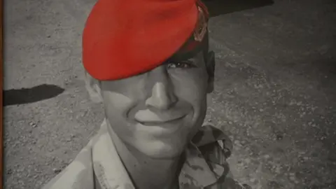 A picture of Corporal Simon Miller shows him smiling and in full uniform. His cap is red.