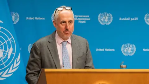 Getty Images Stephane Dujarric speaking at a new conference. He is standing behind a lectern marked with the UN seal and is wearing a grey suit. 