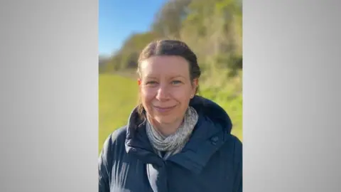 LOUISE THIRLWELL Louise Thirlwall is smiling at the camera. Behind her is a field and trees. 