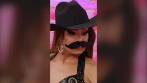 Mariah is wearing a cowboy hat and a fake curly moustache.