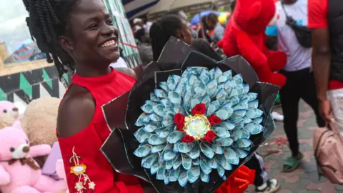 LightRocket/Getty Images A woman in a market in Kenya holding a bouquet made out of blue coloured Kenyan shilling banknotes. Behind her is a stall selling teddy bears.