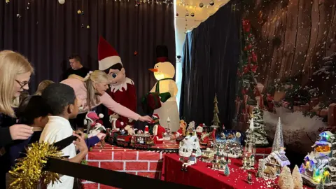 Nicola Haseler/BBC Adults and children stand next to one of the sections which has tables covered in red cloths and winter scenes, including a train. Behind it is a snowy scene with fir trees and snow, and next to it is a large inflatable snowman and Pinocchio.