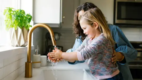 A little girl smiling while washing her hands under the kitchen tap while a woman holds her