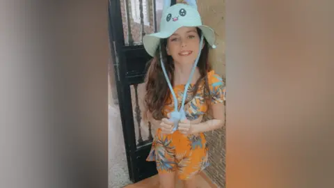 A young girl with long brown hair wearing matching orange shorts and top with a palm tree design on them and a blue summer hat with a cartoon character face on it. She is smiling up at the camera. She is standing in front of a door.