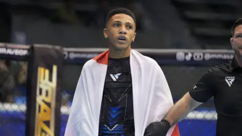 Cairo Thomas pictured in an MMA fighting ring either before or after a competition. He has a white towel wrapped around his shoulders. A coach or a referee stands next to home whilst holding his arm.