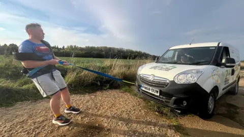 Mike Land pulls a white van on a concrete track full of bumps and bumps, surrounded by green fields and blue skies. He wears shorts, sneakers, a Superman t-shirt, and a pull harness.