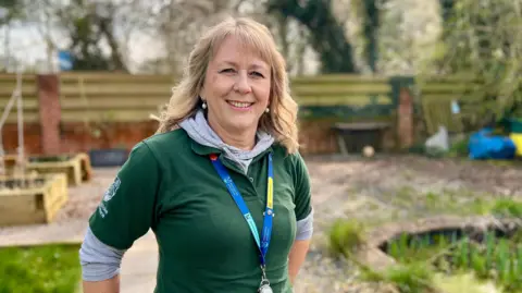 Green Team member Antonia is in the centre of the picture, wearing a green t-shit and a lanyard. She is standing in a garden, with a fence in the background, vegetable planters behind her to her left, and a pond to her right.