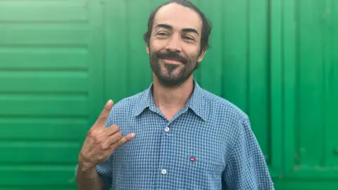 Owen Sennitt/BBC A man with a beard wearing a blue chequered shirt smiles towards the camera in front of a green shed