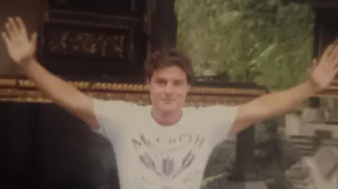 Matthew Osborne is pictured in a grainy image wearing a white t-shirt with a print in the centre. His hands are outstretched