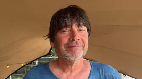 A head and shoulders image of Blur's Alex James. He is wearing a blue T-shirt and smiling while looking into the camera.